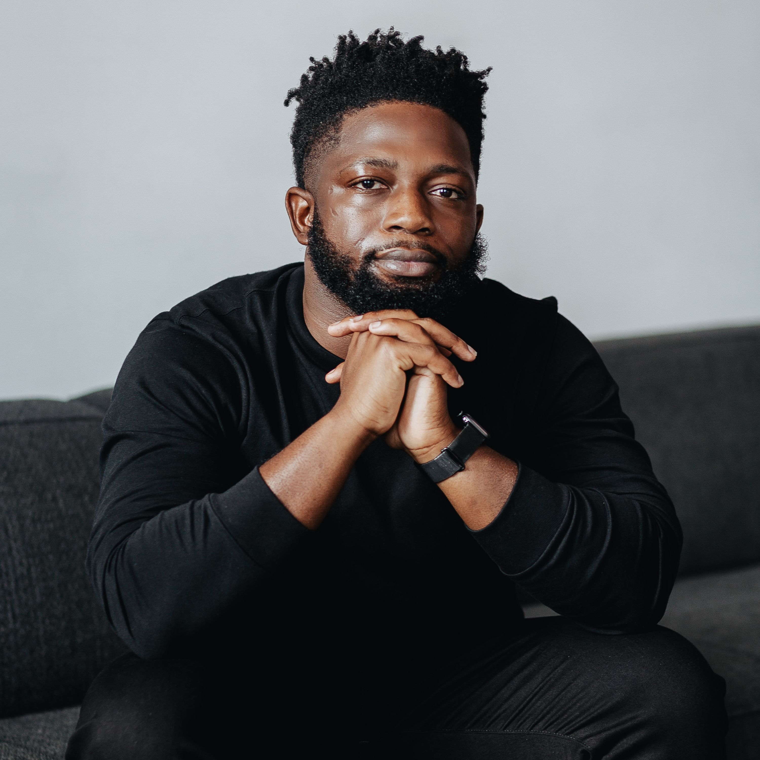 A bearded man in a black sweater sitting with his chin resting on his clasped hands